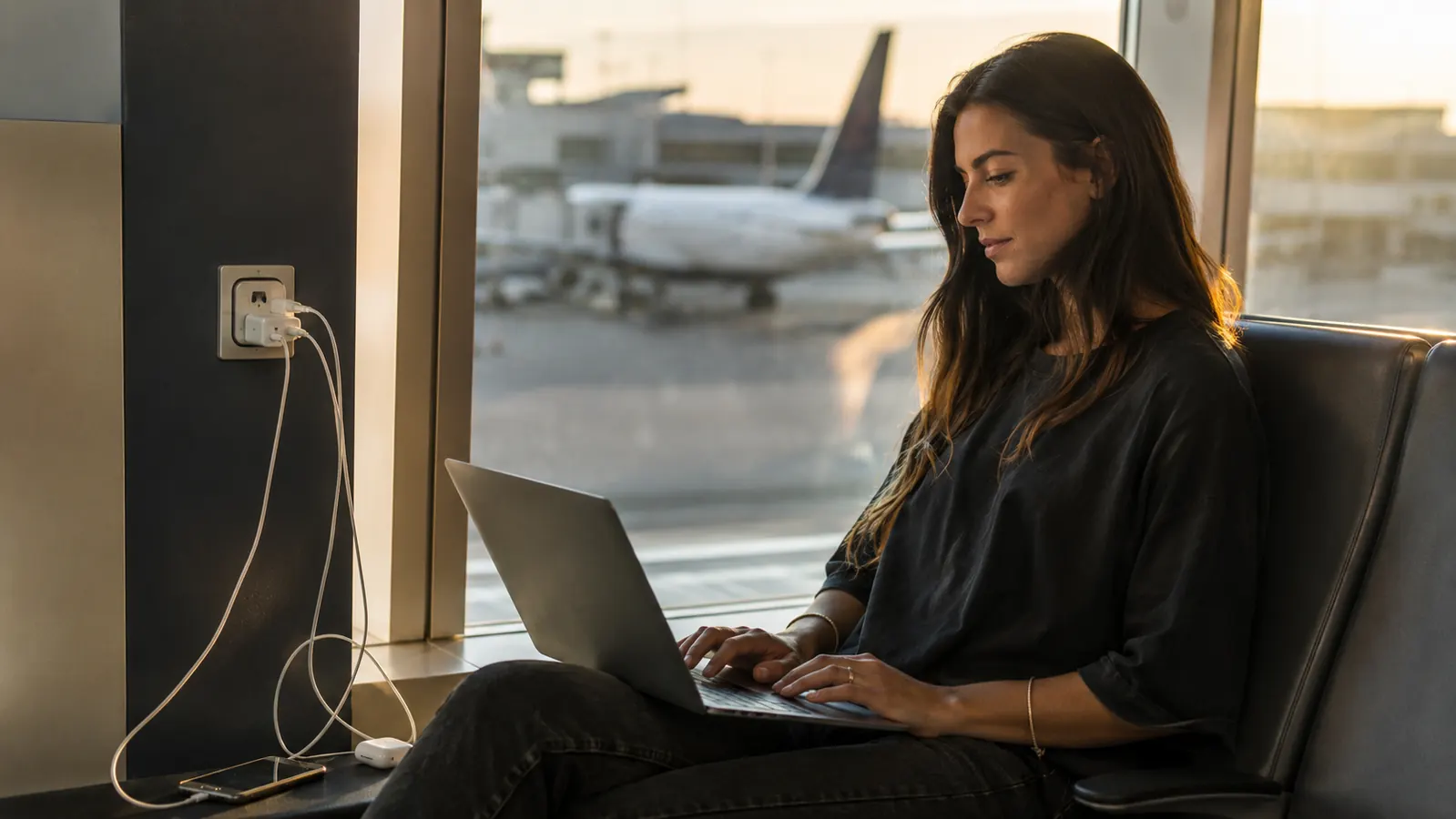 Traveler charging a laptop, phone, and earbuds simultaneously using a universal travel adapter at an airport lounge