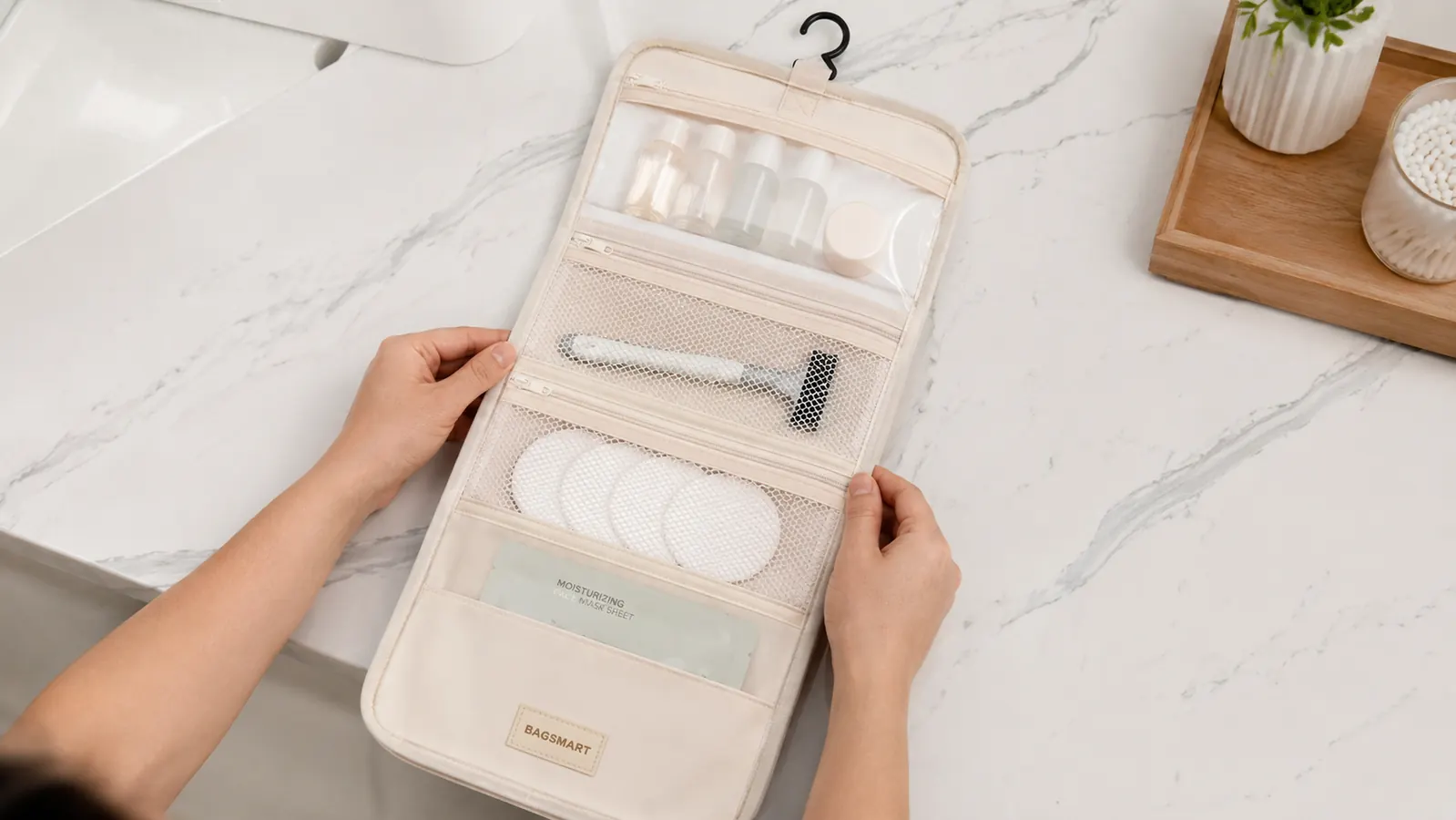 Hanging toiletry bag unfolded flat on a white marble bathroom counter showing all four compartments with travel toiletries sorted by type