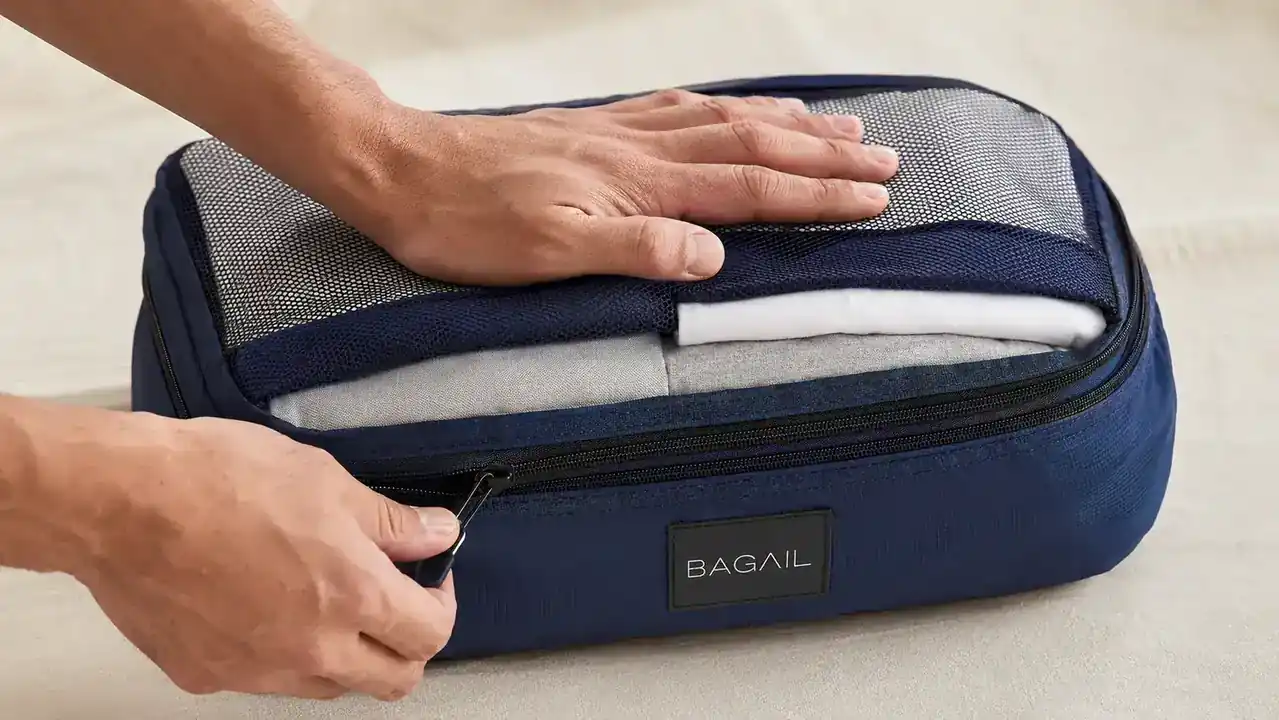 Hands compressing a BAGAIL packing cube zipper closed over a stack of folded clothes